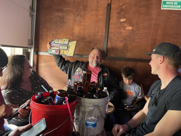 Ted Moncure holds up a trophy in a wood-paneled room with a family sitting around a table with buckets of beer and a water bottle.