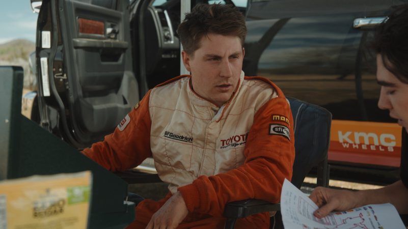 A race car driver in an orange and white firesuit sitting in a chair, talking to a person holding a printed document.
