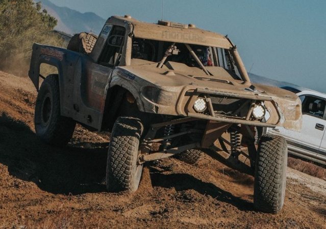 A mud-covered trophy truck driving on a dirt track.