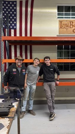 Three men posing in a workshop in front of a hanging American flag and orange metal shelving.