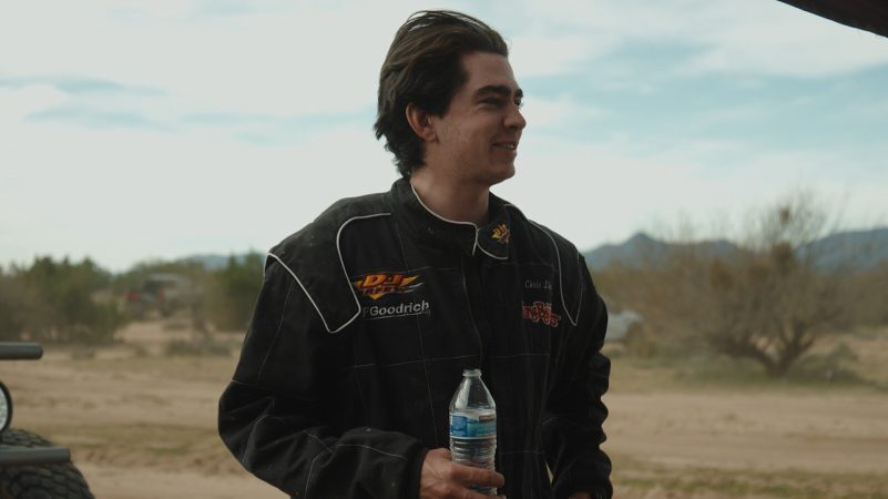A man wearing a black racing suit stands in a desert landscape holding a plastic water bottle.