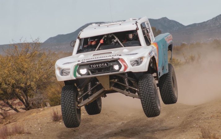 A white Toyota trophy truck jumps over a dirt path in a desert landscape.