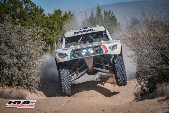 A white Toyota off-road racing truck jumps over a dirt trail in a wooded area.