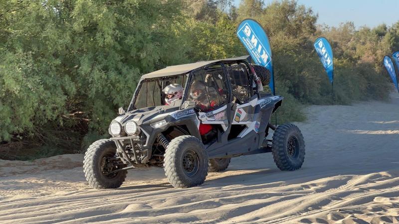 Two people wearing helmets drive a four-seater off-road vehicle along a sandy trail.