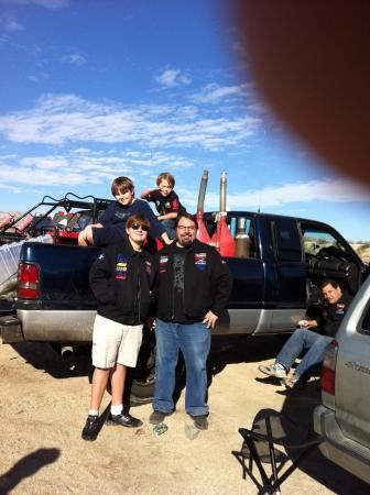 A man and a youth stand in front of a blue pickup truck with two boys sitting on the tailgate and another man seated on the ground nearby.