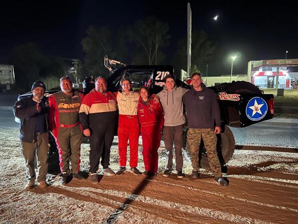 Six people stand in a line at night in front of a parked off-road racing truck numbered 210.