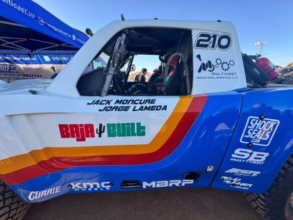 Side view of a white, blue, and yellow off-road racing truck numbered 210, featuring sponsor logos and the names Jack Moncure and Jorge Lameda on the door.