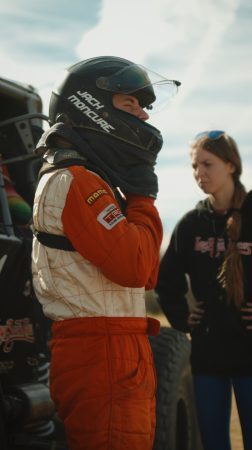 A race car driver in an orange and white firesuit adjusts their helmet while standing next to a woman in a black hoodie.
