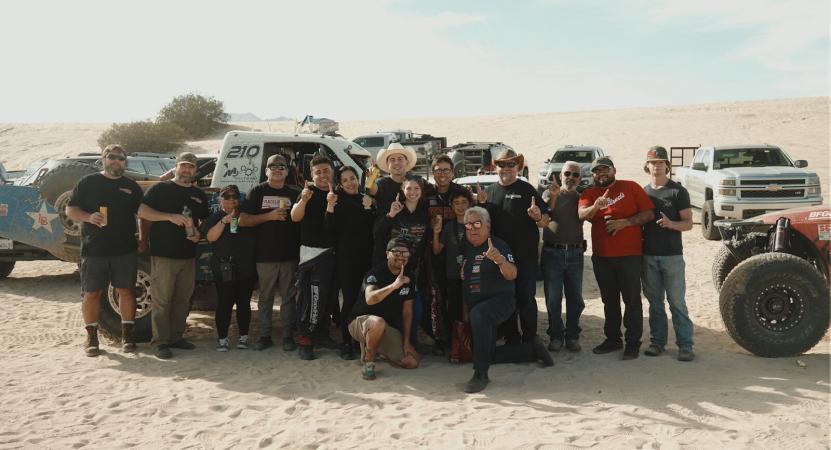 A large group of people posing in a desert landscape in front of off-road racing trucks.