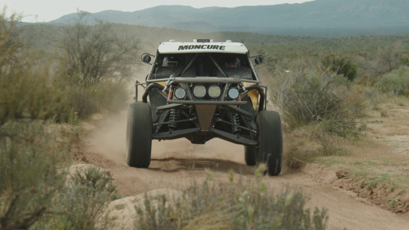 Off-road racing vehicle with the word MONCURE on its roof racing along a dirt trail.