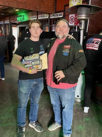 Jack Moncure and Ted Moncure stand together in a bar, with the younger man on the left holding a trophy for a racing competition.