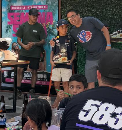 A man in a blue t-shirt puts his arm around a young boy holding a small race car trophy, while another man stands to the left in front of an event banner.