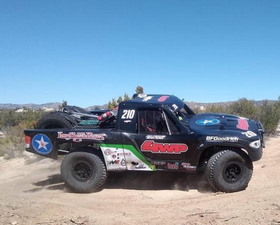 A black off-road racing truck with sponsor decals and the number 210 kicks up dust while driving on a dirt track.