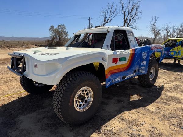 A white and blue off-road trophy truck with Baja Built branding and sponsor decals parked on a dirt lot.