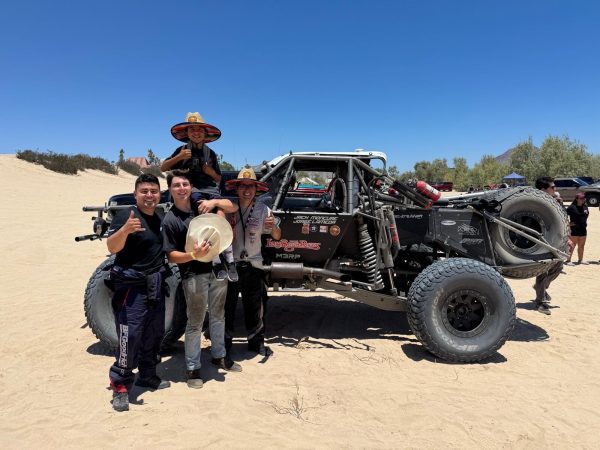 Three men and a child in sun hats pose in front of a specialized off-road racing truck in a desert landscape.