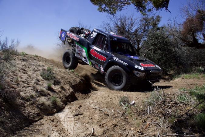 A black off-road racing truck numbered 210 jumps over a dirt trail.