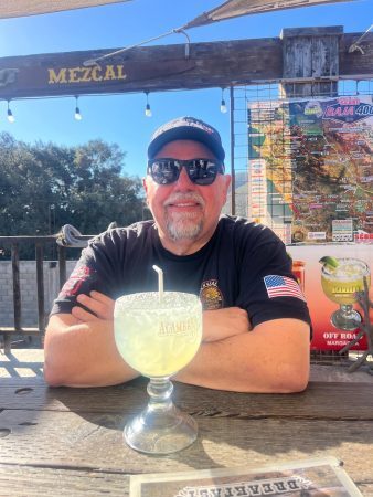 A man wearing sunglasses and a baseball cap sits at an outdoor table with a large margarita in front of him, in front of a Baja 400 race map.