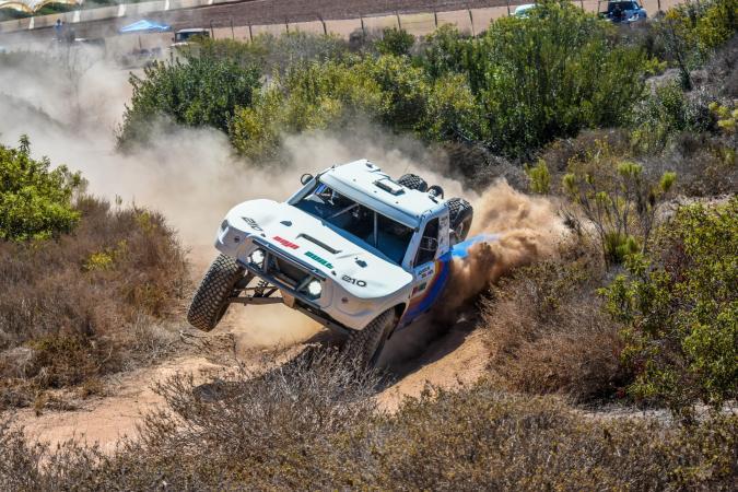 A white off-road racing truck jumps over a dirt trail, kicking up a large cloud of dust.