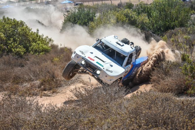 A white off-road racing truck kicks up dust while navigating a turn on a dirt trail.