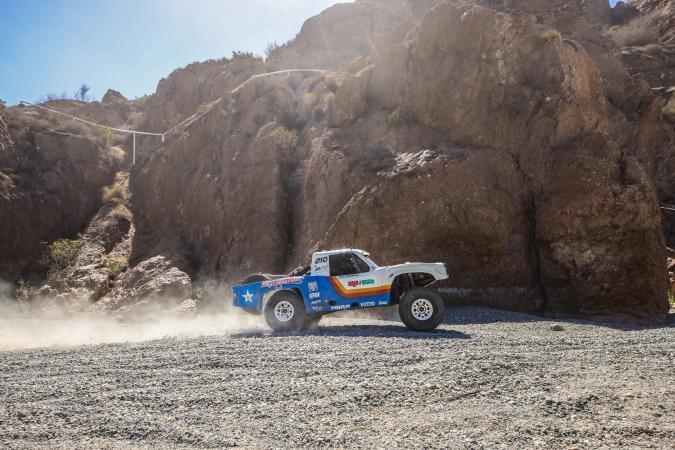 A white and blue off-road racing truck kicks up dust as it drives along a rocky desert track at the base of a cliff.
