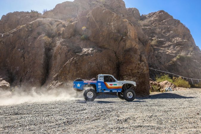 A white and blue off-road racing truck kicking up dust while driving past a large rock formation.