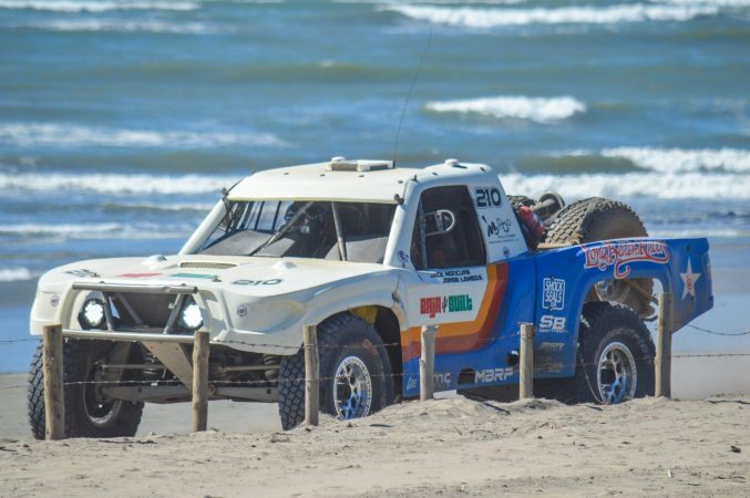A white, orange, and blue racing truck, number 210, drives along a sandy beach with the ocean in the background.