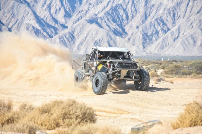 A black Toyota off-road racing truck speeds across a desert track, kicking up a large cloud of dust with mountains in the background.