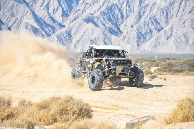 A Toyota off-road racing truck speeds through the desert, kicking up a large cloud of dust.