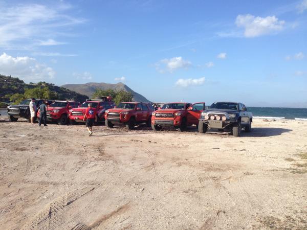 A line of off-road pickup trucks parked on a sandy beach near the ocean.
