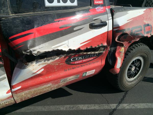 Side view of a red and black off-road truck with severe damage to the driver-side door and rear quarter panel, featuring Certified Used Vehicles branding.