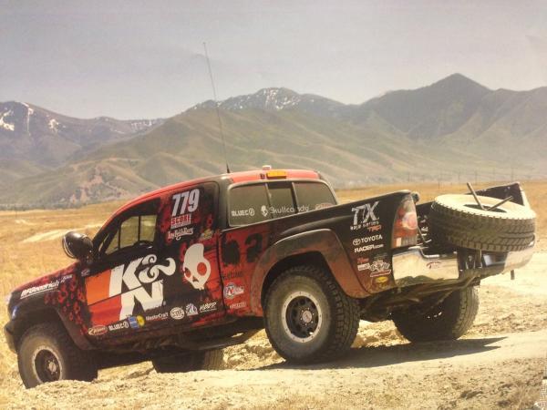 A K&N branded Toyota off-road racing truck with a spare tire mounted in the bed, parked on a dirt trail in front of mountains.