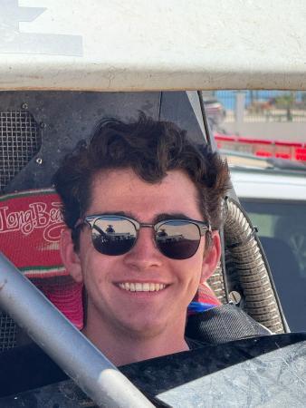 A smiling young man wearing sunglasses sits inside the cockpit of a race car.