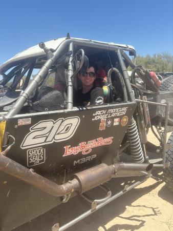 A smiling person wearing sunglasses sits in the drivers seat of a black off-road racing vehicle labeled with the number 210 and Long Beach Racers.