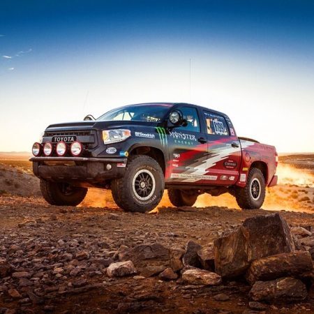 A black Toyota Tundra off-road racing truck with Monster Energy decals driving over rocky desert terrain.