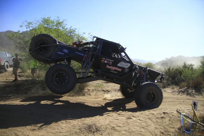 A black off-road race truck, number 210, jumps over a dirt hill on a desert course.