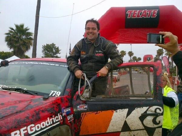 A smiling race car driver stands in the open door of his mud-splattered red and black race truck at an event with an inflatable archway.
