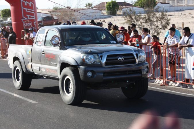 A grey Toyota Tacoma race truck driving past a crowd of spectators standing behind a metal barricade.