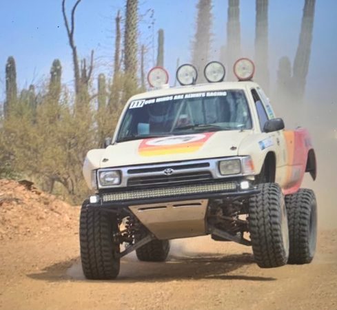 A white off-road racing truck with light bars and racing decals jumps over a dirt path in a desert landscape.