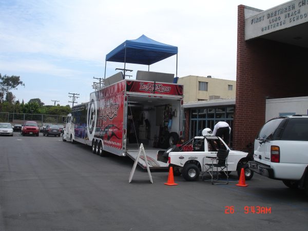 A racing trailer with a blue canopy parked next to a white race car in a church parking lot.