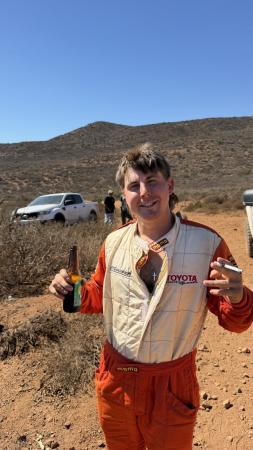 A person wearing an orange and cream racing suit holding a beer bottle and a cigarette in a desert landscape.