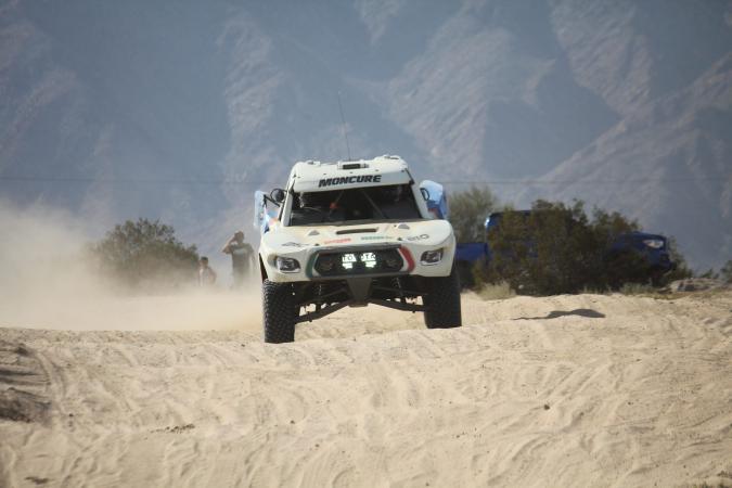 A white Toyota off-road racing truck kicks up dust while driving through a sandy desert landscape.