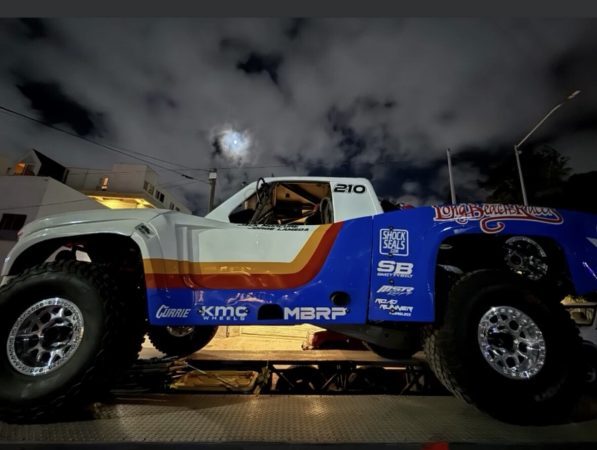 A white, red, and blue trophy truck numbered 210 on a flatbed trailer at night with a full moon in the background.