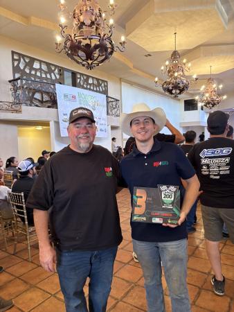 Two men stand together in a hall, one wearing a baseball cap and the other in a cowboy hat holding a La Raza 200 third-place trophy.