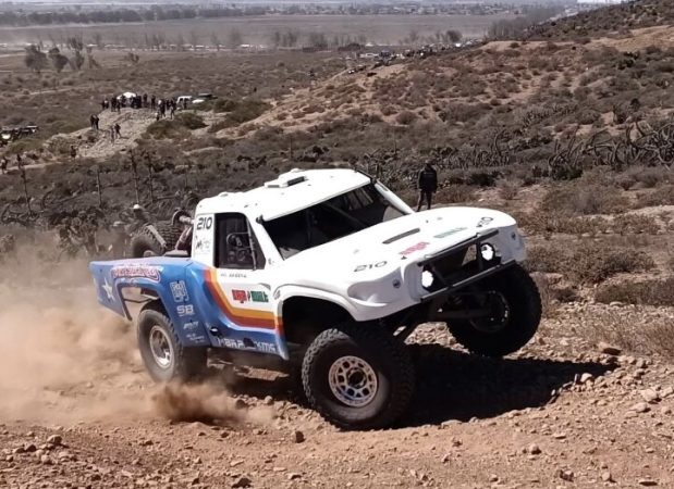 A white off-road racing truck with blue and orange stripes drives up a dirt incline, kicking up dust.