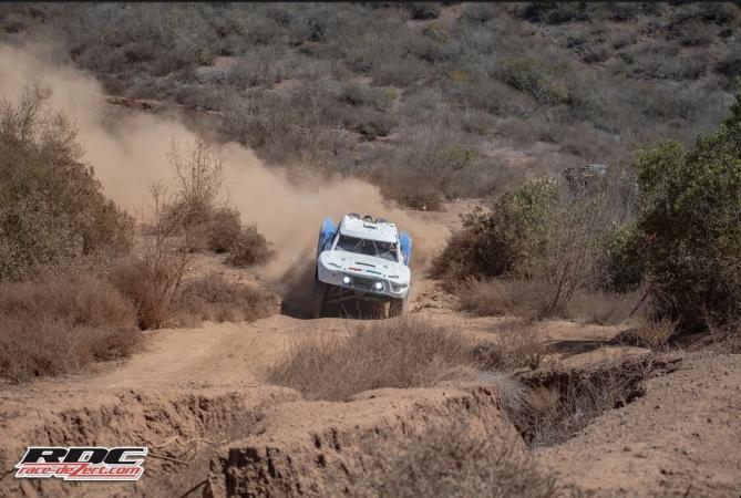 A white off-road racing truck kicks up dust while driving through a desert trail.