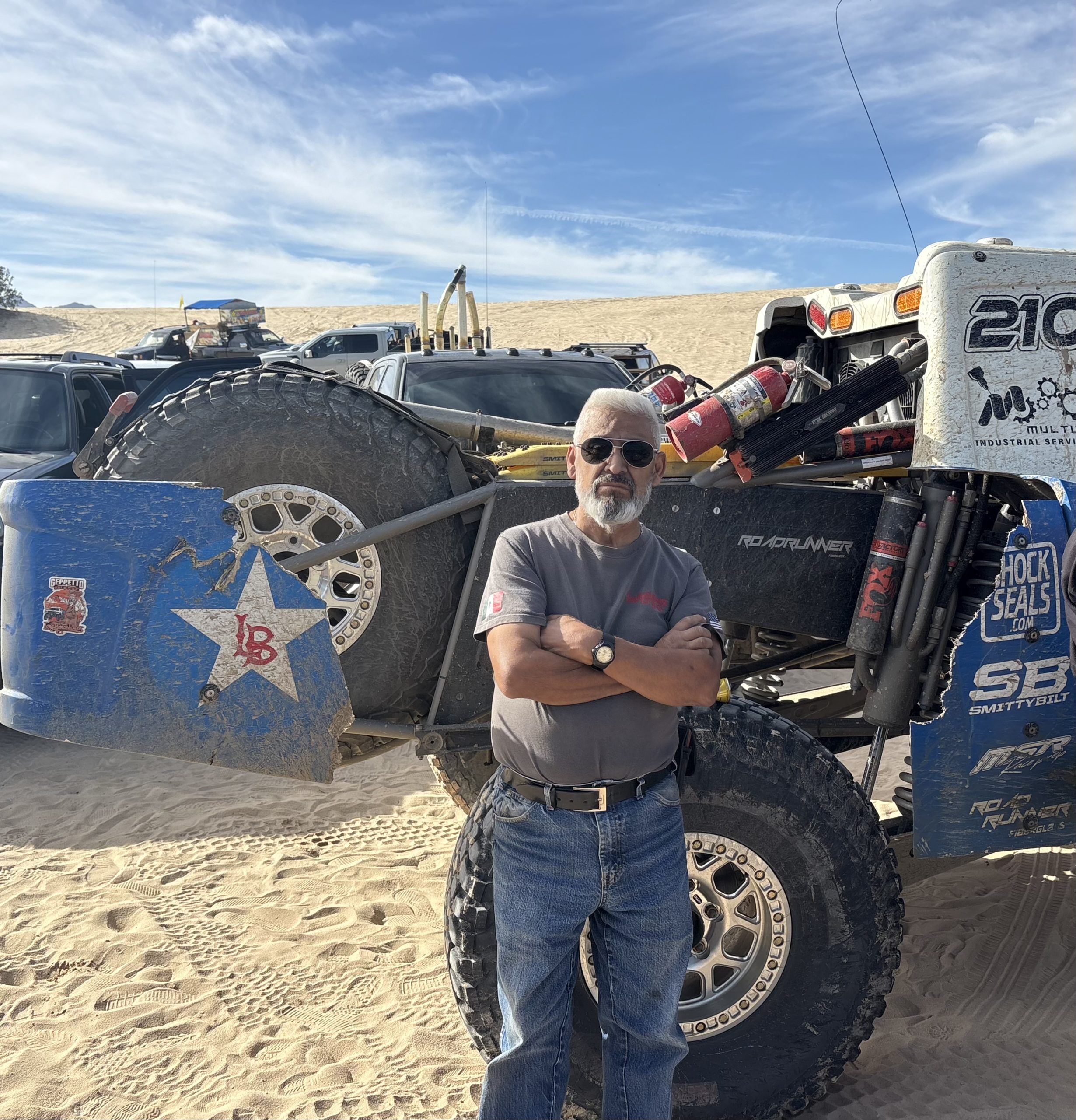 A man with a grey beard and sunglasses stands with his arms crossed in front of a blue and white desert racing truck in a sandy landscape.