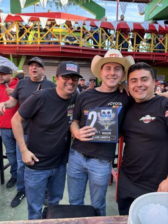 Three men smiling together, with the center man holding a racing trophy that reads Clase Abierta and 30 Aniversario.