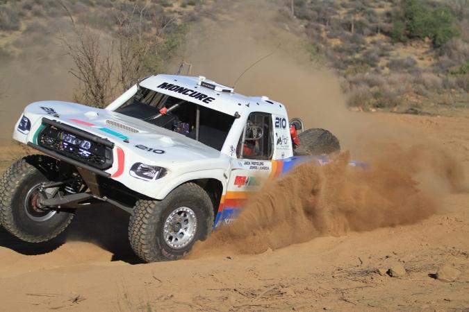 A white off-road racing truck numbered 210 kicks up a large cloud of dust while driving through a desert landscape.