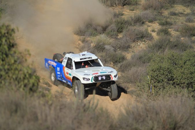 A white, blue, and orange off-road racing truck kicks up dust while driving through a desert trail.