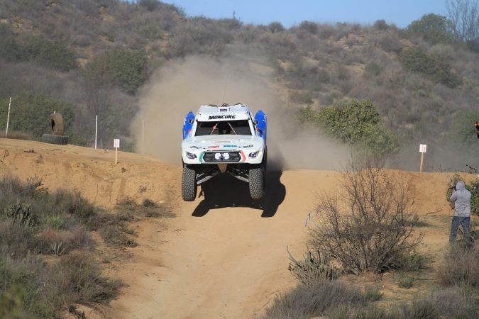 A white off-road racing truck jumps over a dirt hill, kicking up a large cloud of dust.